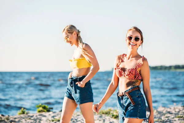 Beautiful smiling young women in sunglasses walking together on sandy beach — Stock Photo