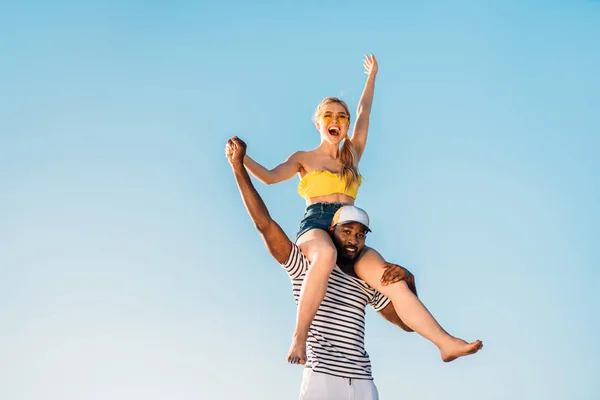 Vista de ángulo bajo de feliz joven pareja multiétnica divirtiéndose juntos contra el cielo azul - foto de stock