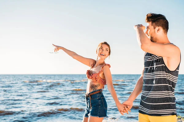 Feliz joven pareja cogida de la mano y mirando hacia otro lado mientras caminan juntos en la playa - foto de stock