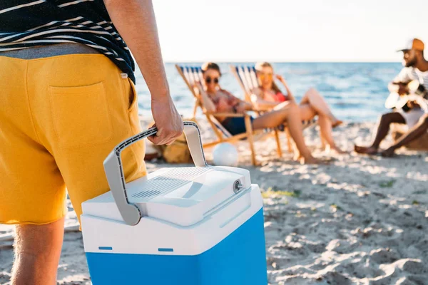 Cropped shot of man holding beach cooler while friends resting on sand behind — Stock Photo