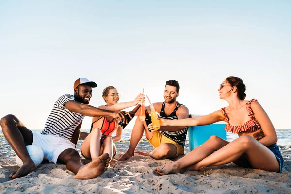 Heureux jeunes amis multiethniques clinking bouteilles en verre avec des boissons tout en étant assis ensemble sur la plage de sable — Photo de stock