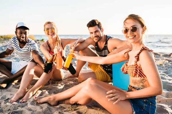 Felices jóvenes amigos multiétnicos sosteniendo botellas de vidrio con bebidas y sonriendo a la cámara en la playa de arena — Stock Photo