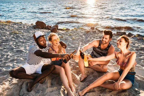 Vista de ángulo alto de felices amigos multiétnicos jóvenes tintineo botellas de vidrio con bebidas en la playa de arena al atardecer - foto de stock