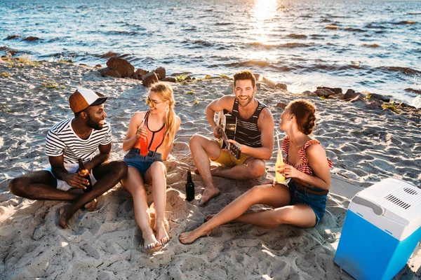 Blick aus der Vogelperspektive auf junge multiethnische Freunde, die bei Sonnenuntergang am Sandstrand Gitarre spielen und Getränke trinken — Stockfoto