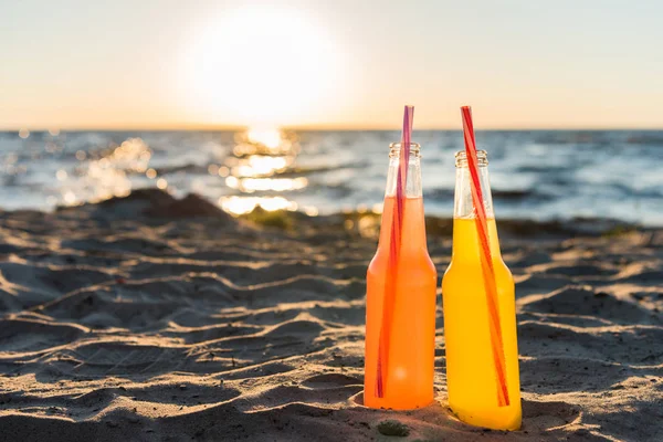 Vista de perto de garrafas de vidro com bebidas refrescantes e palhinhas na praia de areia ao pôr do sol — Fotografia de Stock