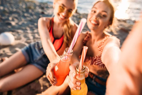 Enfoque selectivo de jóvenes amigas felices sosteniendo botellas de vidrio con bebidas de verano en la playa - foto de stock