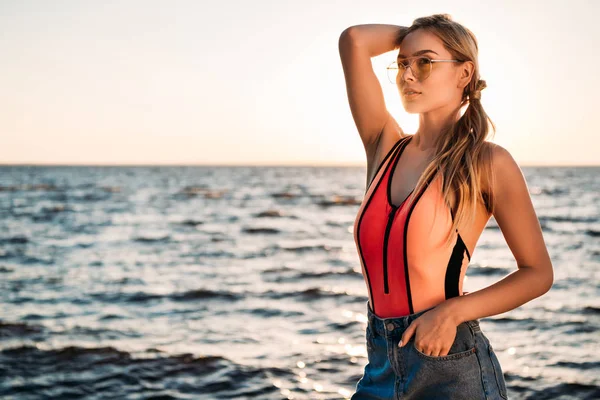 Schöne junge Frau in Sonnenbrille, Badeanzug und Jeanshose, die bei Sonnenuntergang am Strand wegschaut — Stockfoto