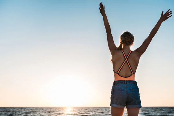 Вид сзади молодой женщины, поднимающей руки и смотрящей на красивый закат над морем — стоковое фото