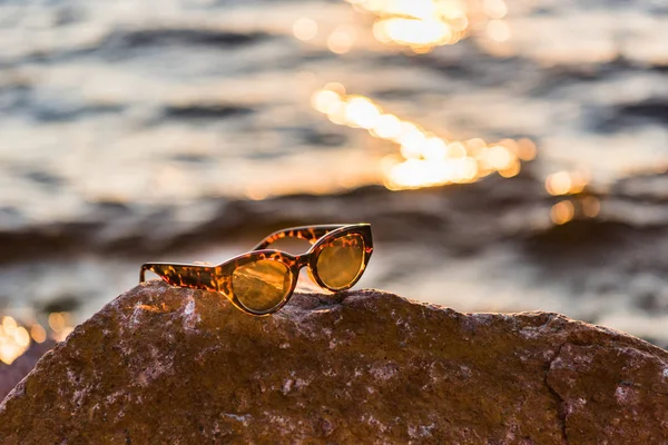 Vista close-up de óculos de sol em pedra na costa do mar ao pôr do sol — Fotografia de Stock