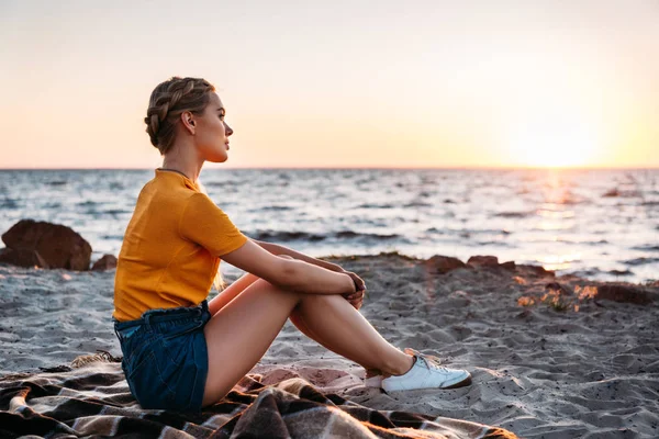 Side view of pensive young woman sitting on plaid at beautiful sea coast at sunset — Stock Photo