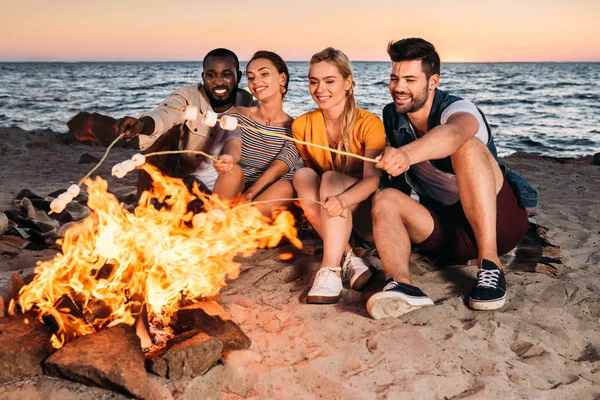 Felices jóvenes amigos multiétnicos asando malvaviscos en la hoguera en la playa de arena al atardecer - foto de stock