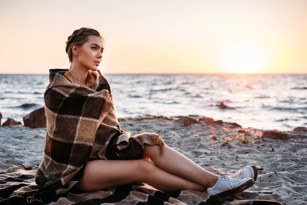 Belle jeune femme coûteuse assise enveloppée de plaid et regardant majestueux coucher de soleil en mer — Photo de stock