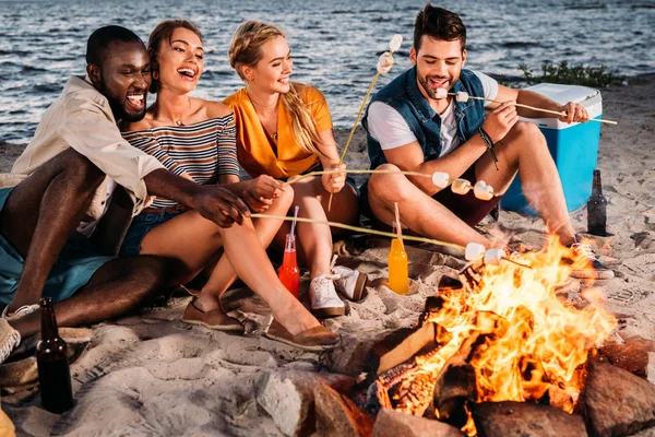 Felices jóvenes amigos multiétnicos asando malvaviscos en la hoguera en la playa al atardecer - foto de stock