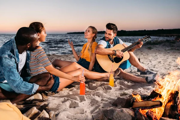 Happy young multiethnic friends enjoying guitar and spending time together on sandy beach at sunset — Stock Photo