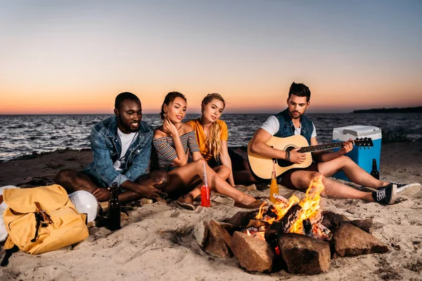 Jovens amigos multiétnicos que gostam de guitarra e passar o tempo juntos na praia de areia ao pôr do sol — Fotografia de Stock
