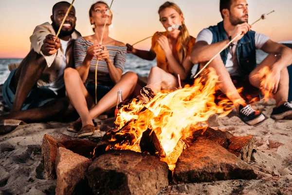 Jeunes amis rôtissant des guimauves au feu de joie tout en étant assis à la plage de sable au coucher du soleil — Stock Photo