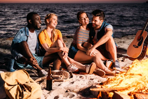 Felizes jovens amigos multiétnicos conversando e passando tempo juntos na praia de areia ao pôr do sol — Fotografia de Stock