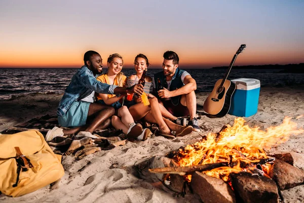 Happy multiethnic friends clinking glass bottles with drinks while spending time together on sandy beach at sunset — Stock Photo