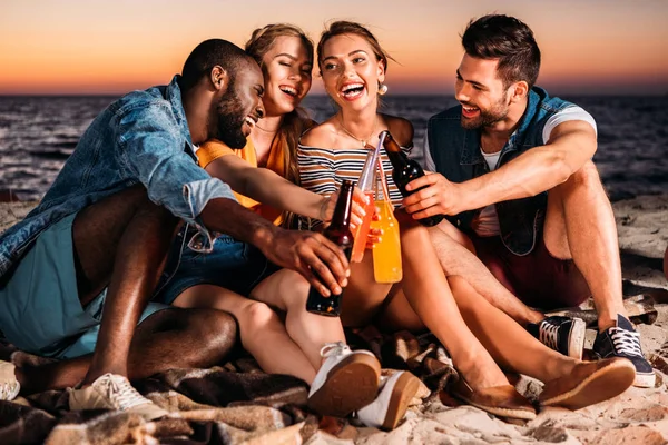 Jovens felizes batendo garrafas de vidro com bebidas enquanto passam o tempo juntos na praia de areia ao pôr do sol — Fotografia de Stock
