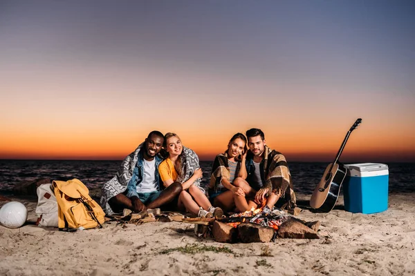 Giovani felici sorridenti davanti alla macchina fotografica seduti insieme sulla spiaggia sabbiosa al tramonto — Foto stock