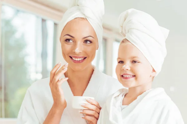 Atractiva sonriente madre e hija en batas de baño y toallas aplicando crema facial juntos - foto de stock