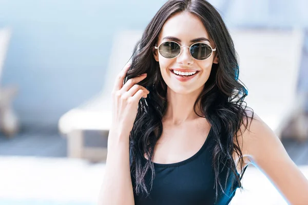 Beautiful brunette girl in swimsuit and sunglasses smiling at camera at poolside — Stock Photo