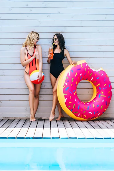 Beautiful smiling young women with beach items drinking summer beverages at poolside — Stock Photo
