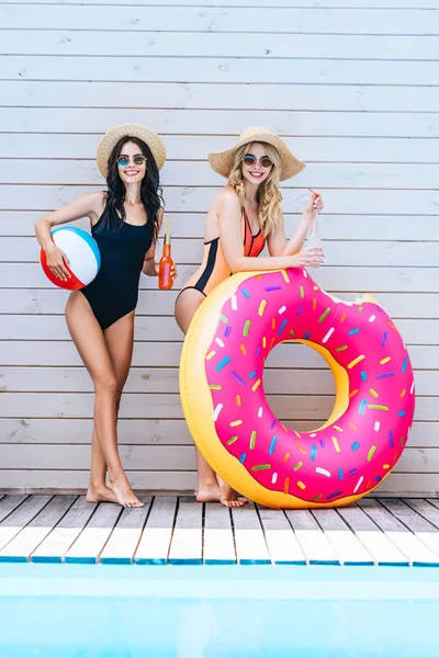 Schöne Mädchen halten Flaschen mit erfrischenden Getränken und Strandutensilien in der Nähe des Swimmingpools und lächeln in die Kamera — Stockfoto