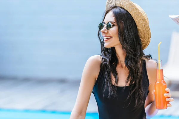 Jeune femme souriante en maillot de bain tenant bouteille avec boisson rafraîchissante et regardant loin de la piscine — Photo de stock