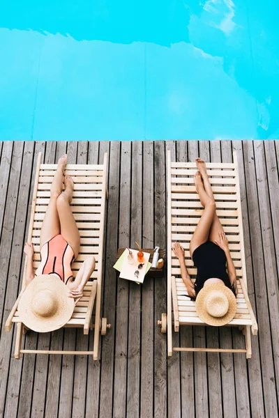 Vista de alto ângulo de mulheres jovens em trajes de banho e chapéus deitados em salões de chaise perto da piscina — Fotografia de Stock