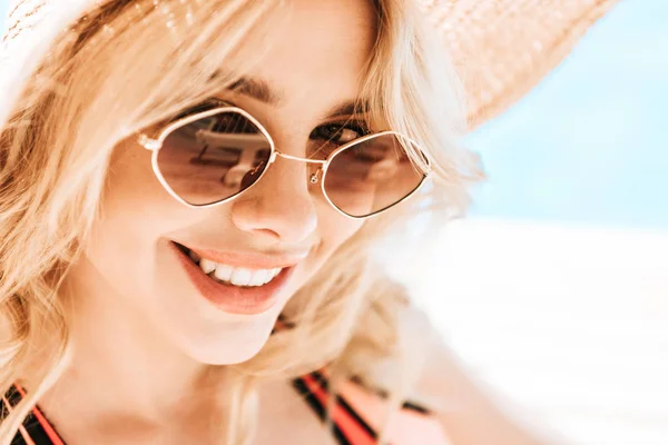 Portrait de belle jeune femme blonde en lunettes de soleil et chapeau en osier souriant à la caméra au bord de la piscine — Photo de stock