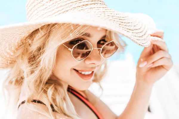 Portrait de belle jeune femme blonde en osier chapeau et lunettes de soleil souriant à la caméra près de la piscine — Photo de stock