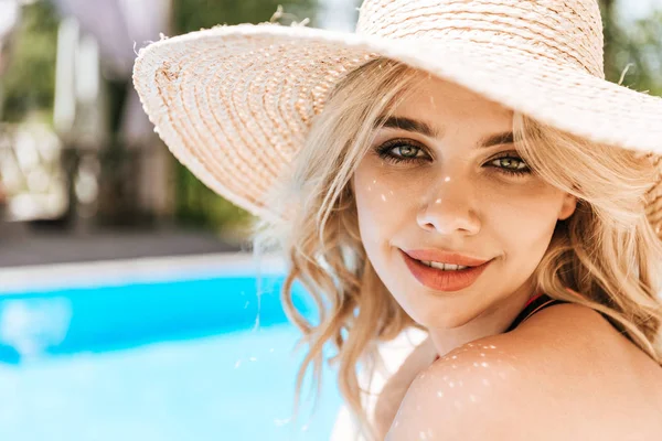 Retrato de hermosa joven rubia en sombrero de paja sonriendo a la cámara cerca de la piscina - foto de stock