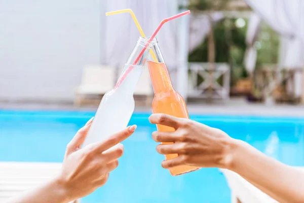 Primer plano vista parcial de las mujeres jóvenes tintineando botellas de bebidas de verano mientras descansan en la piscina - foto de stock