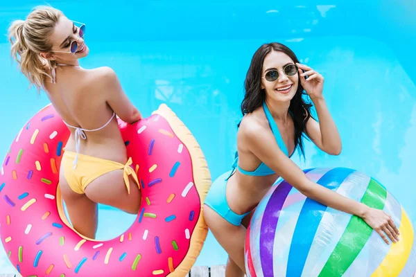 Vista de alto ângulo de mulheres jovens felizes em biquíni e óculos de sol segurando bola inflável e donut na piscina — Fotografia de Stock