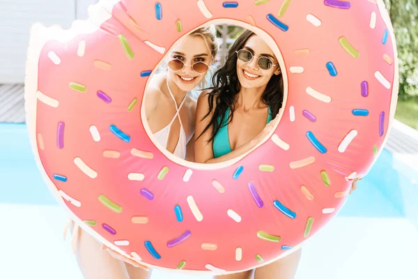 Jeunes femmes heureuses en lunettes de soleil et maillots de bain tenant un beignet de bain et souriant à la caméra près de la piscine — Photo de stock