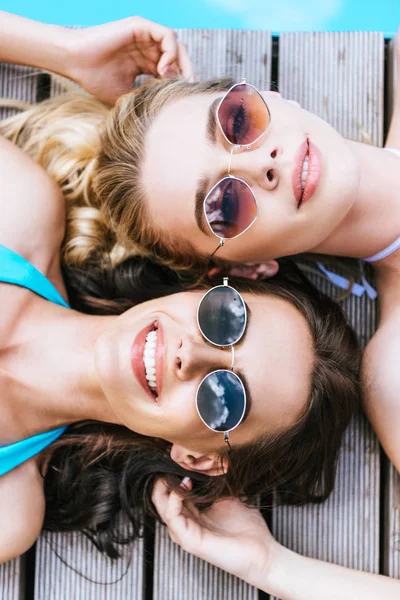 Vue de dessus de belles jeunes femmes en lunettes de soleil souriant à la caméra tout en étant couché au bord de la piscine — Photo de stock