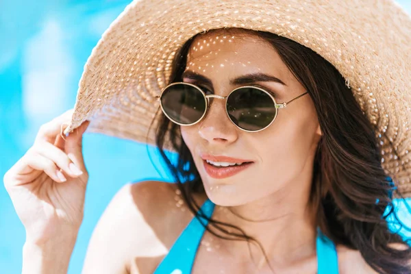Beautiful brunette woman in sunglasses and straw hat smiling at poolside — Stock Photo