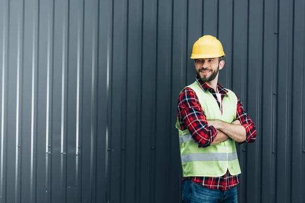 Male worker in safety vest and helmet standing with crossed arms near wall — Stock Photo