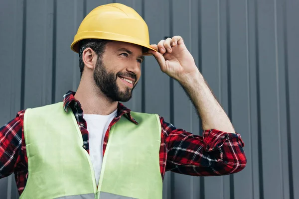 Constructor masculino sonriente en chaleco de seguridad y casco de pie cerca de la pared - foto de stock