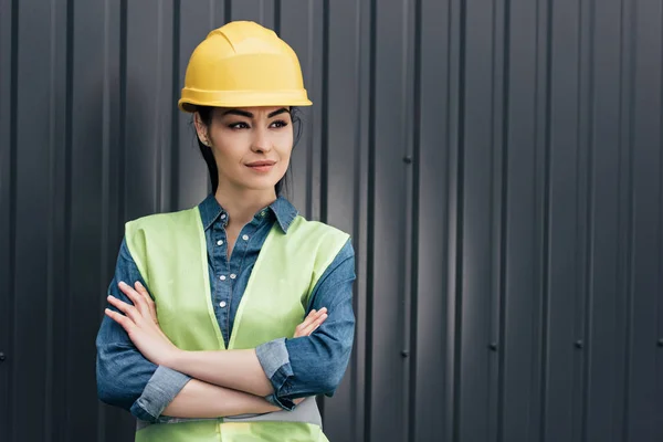 Female engineer in safety vest and hardhat standing with crossed arms at wall — Stock Photo