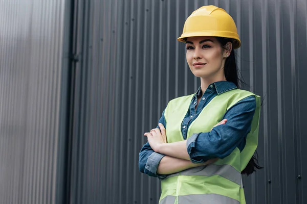 Confident female architect in safety vest and helmet standing with crossed arms at wall — Stock Photo