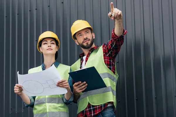 Arquitectos en cascos apuntando y trabajando con planos y portapapeles en el techo - foto de stock