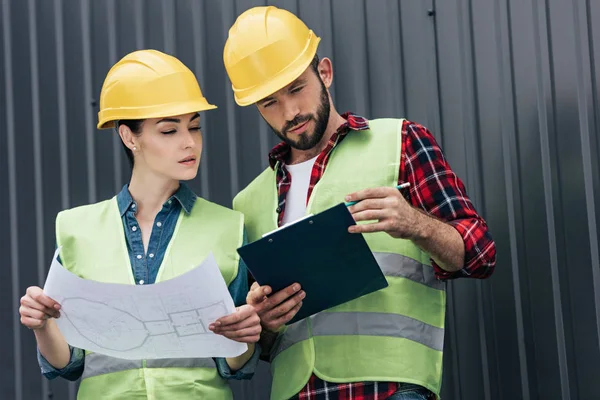 Adult architects in hardhats working with blueprint and clipboard on roof — Stock Photo