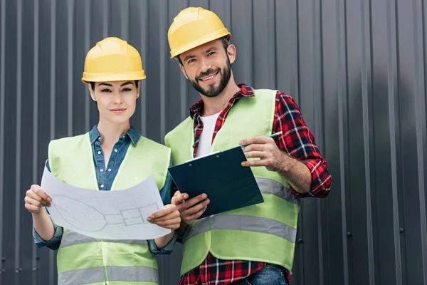 Architectes en gilets de sécurité et casquettes de travail avec plan et presse-papiers sur le toit — Photo de stock