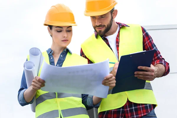 Professional engineers in safety vests and helmets working with blueprints and clipboard on roof — Stock Photo