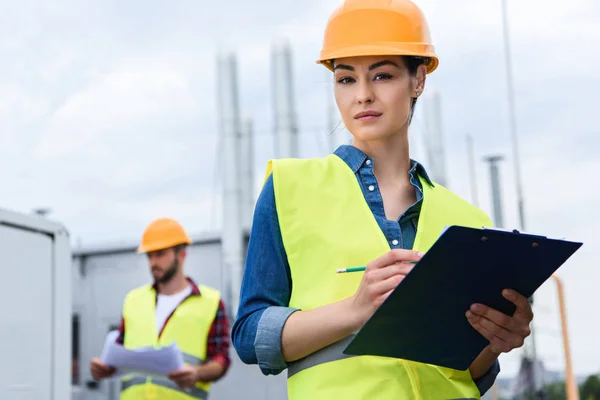 Professional female engineer in helmet writing in clipboard on roof, male colleague with blueprint behind — Stock Photo