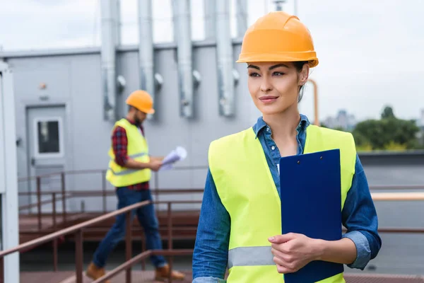 Ingeniero femenino con portapapeles en el techo, colega masculino con plano detrás - foto de stock
