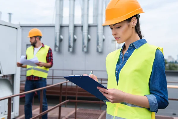 Female architect in helmet writing in clipboard on roof, male colleague with blueprint behind — Stock Photo