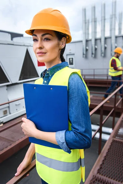 Arquitecto femenino en casco sujetando portapapeles en el techo, colega masculino detrás - foto de stock
